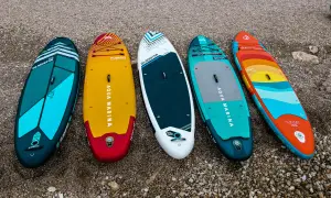 Pięć desek SUP w różnych kolorach i wzorach leży na kamienistej plaży.