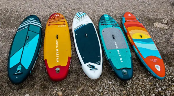 Pięć desek SUP w różnych kolorach i wzorach leży na kamienistej plaży.